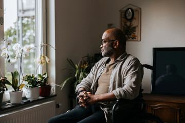 Thoughtful senior man sitting with hands clasped on wheelchair near window at nursing home