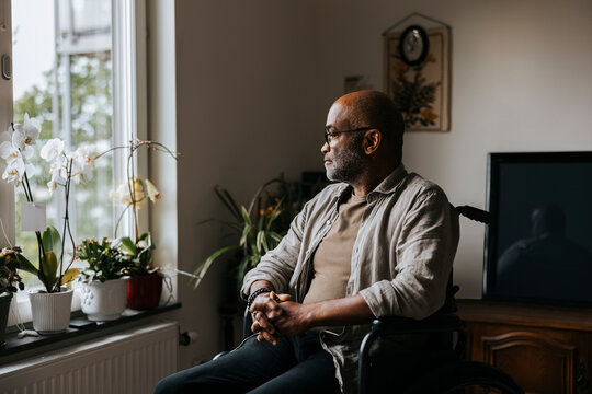 Thoughtful senior man sitting with hands clasped on wheelchair near window at nursing home