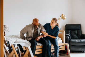 Female caretaker assisting senior man while getting up from bed at nursing home