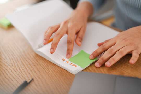 A close-up of a person's hands placing a green sticky note in a notebook with handwritten "work to do" text, on a wooden desk with a laptop in a bright workspace.