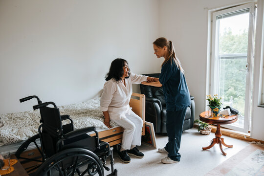 Side view of female nurse assisting senior woman while getting up from bed at nursing home