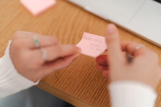 A close-up of a person holding a pink sticky note with a handwritten meeting reminder, with a wooden desk and a laptop in the background in a modern workspace.