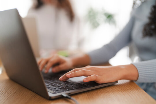 A close-up of a woman's hands typing on a laptop keyboard while working at a wooden desk in a modern office, with another person visible in the blurred background.