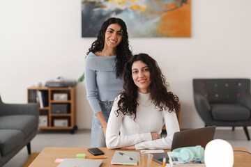 Two young women with curly hair are posing together at a wooden desk in a bright and modern workspace, smiling with laptops, notebooks, and office supplies around them.