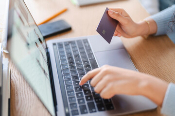 A close-up of a person holding a credit card while using a laptop for online shopping, with a blurred screen displaying clothing items in a modern workspace.