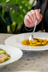A close-up of a hand holding a spoon, about to scoop creamy soup with herbs from a white bowl. In the foreground, a fresh salad and a pastry are visible on a textured table.