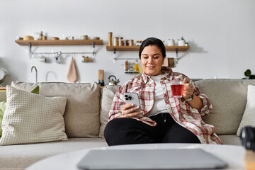 A young plus sized woman relaxes on a sofa, sipping tea while checking her smartphone.