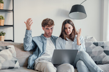 Millennial couple annoyed by stuck laptop or online news sitting together on sofa, frustrated young...
