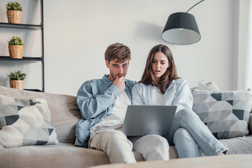 Millennial couple annoyed by stuck laptop or online news sitting together on sofa, frustrated young...