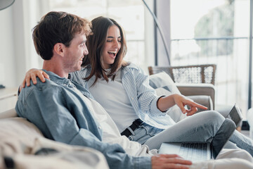 Happy millennial couple sit relax on couch in living room watching video on laptop together, smiling young husband and wife rest on sofa at home browsing Internet using modern computer device.