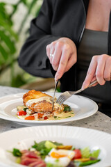 A close-up of hands cutting into a delicious chicken dish on a white plate, surrounded by fresh vegetables. In the foreground, a salad adds color to the dining scene.