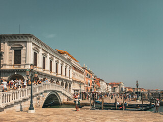 A Sunny Day in Venice. A bridge crosses a bustling canal, lined with colorful buildings and dotted with boats. The sky is a brilliant blue, adding to the cheerful atmosphere.