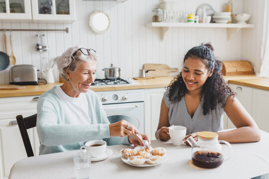 Two females of diverse age and ethnicity having nice conversation during sunday breakfast in cozy bright kitchen, african american young female drinking tea with senior caucasian lady at table