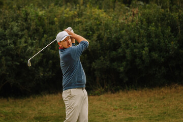 A male golfer in a white cap and blue sweater takes a powerful swing with a golf club, surrounded by greenery on a golf course.