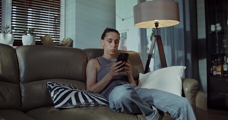 Fototapeta premium A woman uses a mobile phone while sitting on the couch at home. Does she look sad or offended