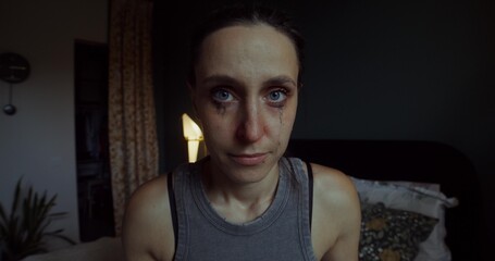 A woman is crying sitting on a bed at home. Her mascara was leaking. She's looking at the camera