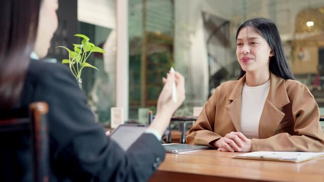 A woman in a brown jacket is sitting at a table with another woman. The woman in the brown jacket is smiling and shaking hands with the other woman
