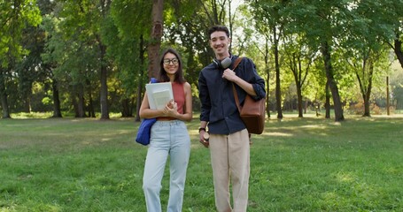 Fototapeta premium A young man and a woman with backpacks and notebooks in their hands are smiling at the camera. They are standing in the city park on a summer day