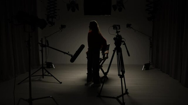 Studio for photo video production with lighting equipment and cyclorama, model actress sits on director chair in the middle under spotlight in front of camera.