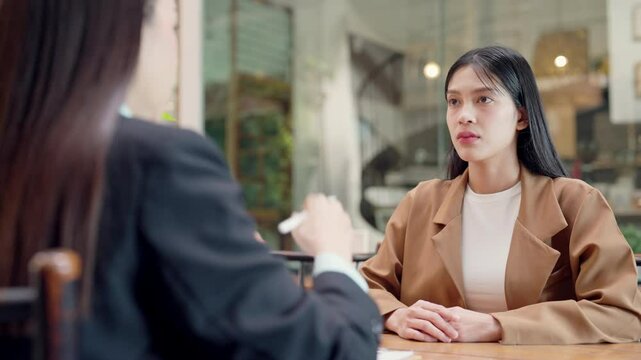 Two women are sitting at a table, one of them is wearing a brown jacket. They are having a conversation