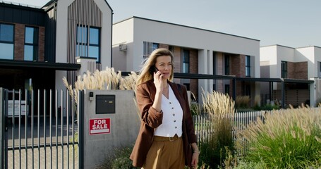 A realtor is talking on a mobile phone standing near a house with a sign for sale. She smiles