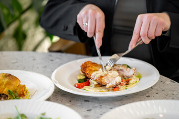 A close-up of hands using a knife and fork to cut into a plated chicken dish with colorful vegetables. The setting is a stylish dining environment, enhancing the culinary experience.