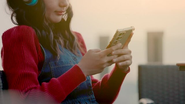 Young smiling asian woman in red sweater and wireless headphones resting with smartphone, enjoying music playlist for Relax and enjoying a peaceful moment outdoors at sunset.