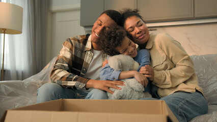 Little kid boy adopted child open cardboard box parcel surprise gift birthday present toy excited happy son hugging teddy bear parents children African American bonding love family at home moving day