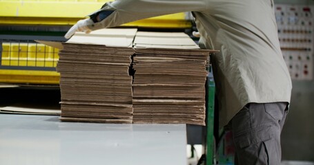 A man in a hard hat works at a paper mill. He picks up the machine-cut cardboard with a concentrated look