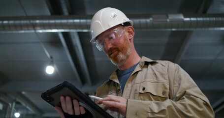 A man in a protective helmet and glasses uses a tablet while working at a factory. He stands near the production machine © m-art