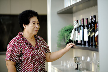 Elderly woman in red patterned blouse standing near home bar, browsing collection of wine bottles on modern black shelf with glassware and decorative plants in well-lit kitchen