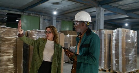 A worker and a boss are talking standing in a paper mill warehouse. A man uses a tablet