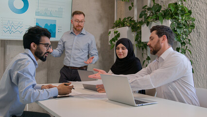 Male leader speaker chief executive talk to diverse multiracial team business people investors businessmen and businesswoman discussion statistic graphs diagrams marketing sales presentation at office