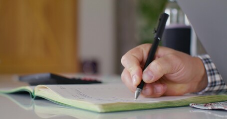 A man writes in a notebook while working. An open laptop on the table. Close-up of his hand, an unrecognizable person