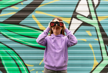 young smiling woman in lavender hoodie observes through binoculars with red lenses, standing in front of colorful graffiti wall. urban setting enhances theme of exploration, curiosity, and discovery