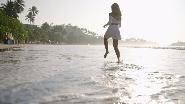 Sporty girl running barefoot in sea surf waves. Young lady in mini skirt jumping along sandy ocean coast. Attractive woman strolling and enjoying sunset at the seaside on vacation. Low angle.