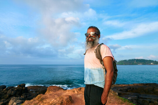 Elderly man with backpack hikes by ocean coast. Senior with gray beard enjoys nature travel. Adventurous journey in retirement. Happy explorer with active lifestyle, young spirit embraces scenic view.
