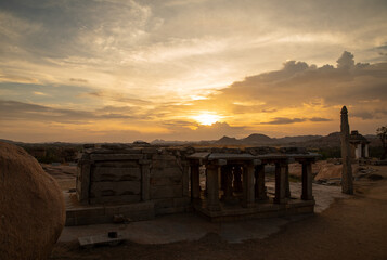 The ancient stone temples of Hampi stand in serene grandeur against a dramatic sunset, reflecting...
