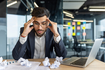 Frustrated and overwhelmed man sitting at workplace inside office, unsuccessful financial report....