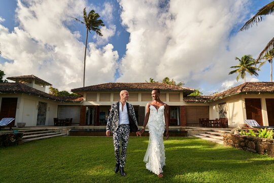 Ambiguous gender fluid person in white bridal dress and lgbtq groom in bold suit walk hand in hand at tropical villa wedding. Diverse love, unique celebration, inclusion moments. - Powered by Adobe