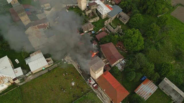 Firefighters extinguish a fire in Svan tower, Mestia. Smoke and flames visible. Fire trucks on-site, people watching. Rural village setting with old stone buildings, green fields. Aerial shots. Drone