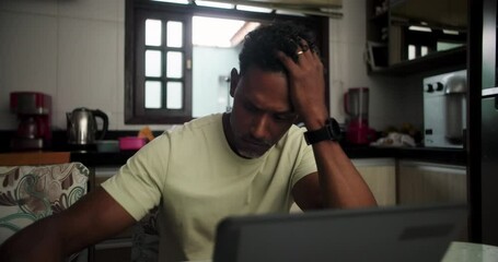 Man of African descent sitting at kitchen table, hands gripping head, deeply stressed, frustrated, looking down at laptop, struggling with work, financial problems, mental burden