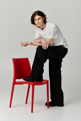 A handsome young man showcases his unique fashion style while leaning on a vibrant red chair.