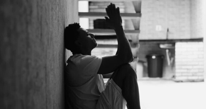 Man of African descent sitting on ground against wall, arms stretched forward, lost in thought, emotional exhaustion, deep personal reflection, struggling with inner turmoil