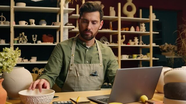 Caucasian salesman seller potter male man guy businessman artist small business owner examining bowl clay ceramic writing notes working new design laptop computer device inside pottery workshop store