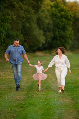 Naklejka premium Happy parents having fun walking together in park in summer. Mom, dad, daughter having fun together running together in meadow holding hands smiling happily. Happy family, parenthood concept. Vertical