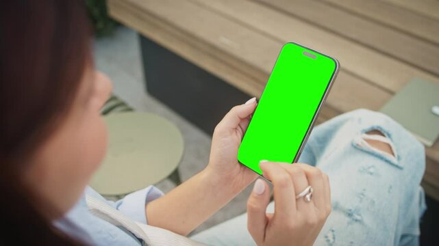 Person holding smartphone with green screen while sitting outdoors on wooden bench. Fingers interacting with touchscreen. Scrolling or zooming. Notebook and wireless earbuds on table. Chroma key.