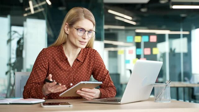 Businesswoman watching video call online conference taking notes looking at laptop computer screen sitting in office.  Woman listening remote business training or course talking with a tutor