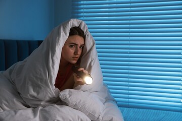 Fear of darkness. Scared young with blanket and flashlight on bed indoors at night, space for text