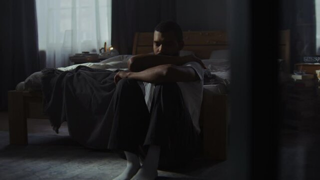 Frustrated black man sits on floor hugging shoulders at home